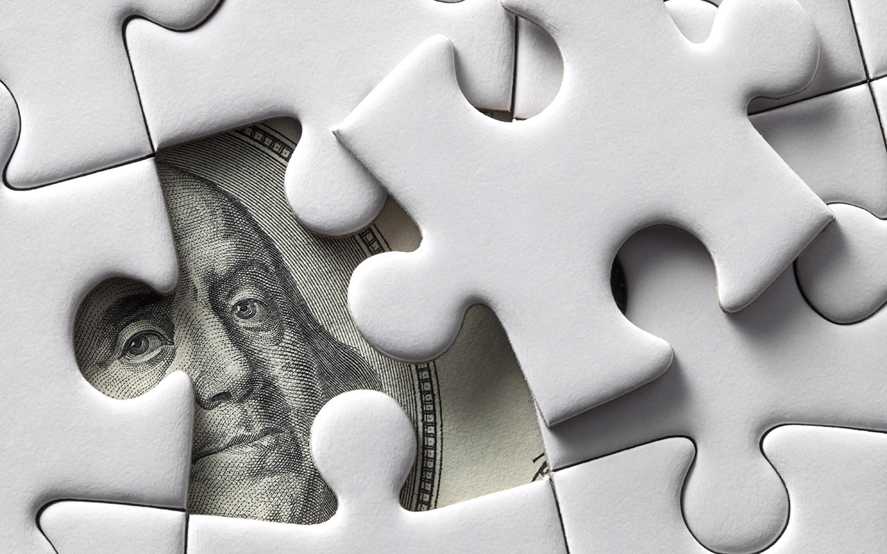 Photo of a puzzle piece being placed into the correct spot in the puzzle, behind which is a $100 bill